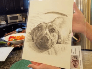 Person holding a detailed pencil drawing of a dog on a countertop, with a bowl of fruit in the background.
