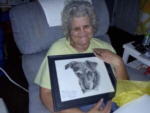 Elderly woman sitting in a chair, smiling and holding a framed black-and-white drawing of a dog.