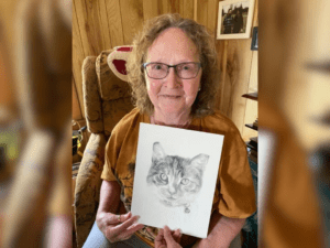 A woman with glasses sits on a chair holding a drawing of a cat. She is smiling, and the setting has wooden walls and framed pictures.