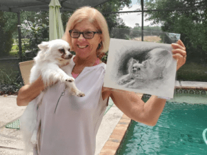 A woman holding a small white dog and a pencil sketch of the same dog, standing beside a pool in an outdoor setting.
