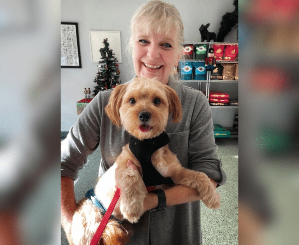 Linda holds a small, light brown dog on a red leash in a room with a Christmas tree and colorful items on shelves.